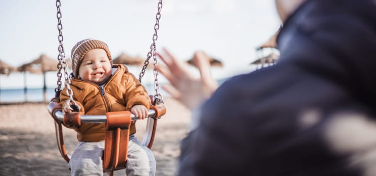 Mother pushing her cheerful infant baby boy child on a swing on sandy beach playground outdoors on nice sunny cold winter day in Malaga, Spain