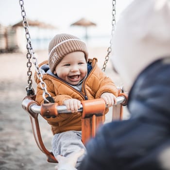 Mother pushing her cheerful infant baby boy child on a swing on sandy beach playground outdoors on nice sunny cold winter day in Malaga, Spain