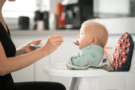 Mother spoon feeding her baby boy child in baby chair with fruit puree in kitchen at home. Baby solid food introduction concept