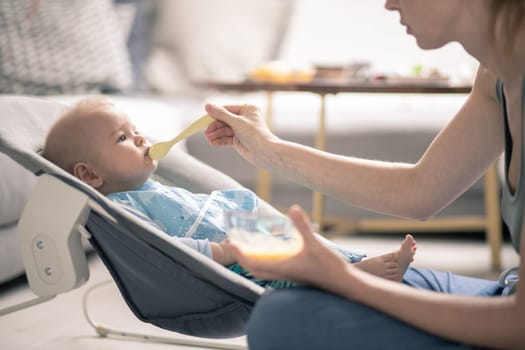 Mother spoon feeding her baby boy child in baby chair with fruit puree. Baby solid food introduction concept