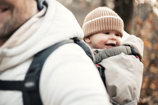 Sporty father carrying his infant son wearing winter jumpsuit and cap in backpack carrier hiking in autumn forest