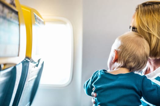 Mom and child flying by plane. Mother holding and playing with her infant baby boy child in her lap during economy comercial flight. Concept photo of air travel with baby. Real people