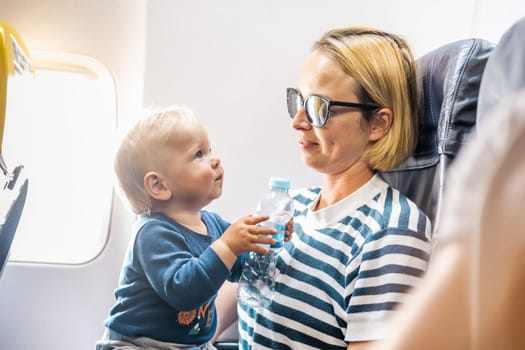 Mom and child flying by plane. Mother holding and playing with her infant baby boy child in her lap during economy comercial flight. Concept photo of air travel with baby. Real people