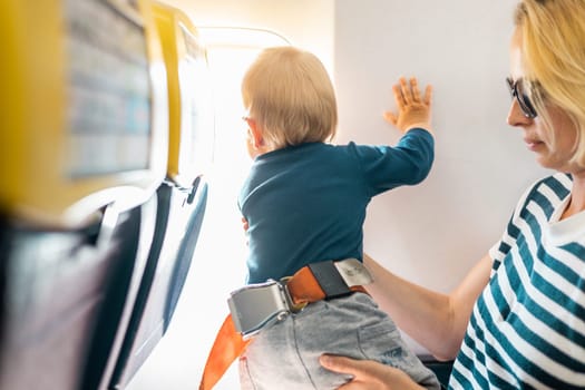 Mom and child flying by plane. Mother holding and playing with her infant baby boy child in her lap during economy comercial flight. Concept photo of air travel with baby. Real people
