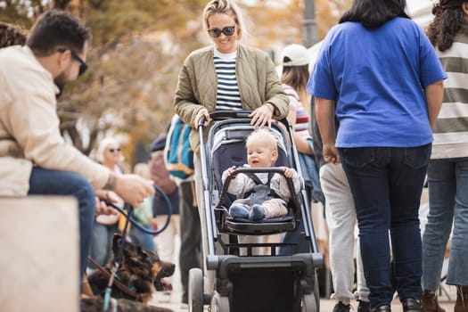 Mother waling and pushing his infant baby boy child in stroller in crowd of people wisiting sunday flea market in Malaga, Spain