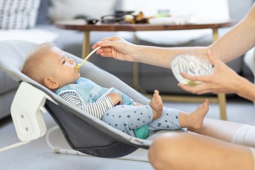 Mother spoon feeding her baby boy child in baby chair with fruit puree. Baby solid food introduction concept
