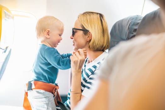 Mom and child flying by plane. Mother holding and playing with her infant baby boy child in her lap during economy comercial flight. Concept photo of air travel with baby. Real people