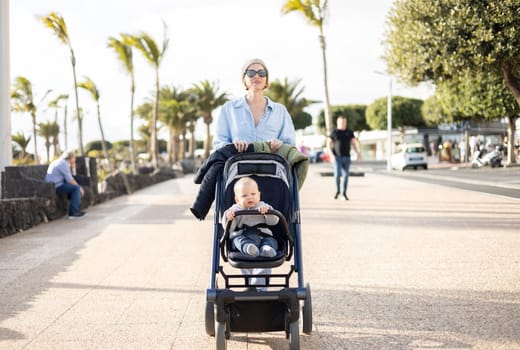 Fashinable mother walking and pushing her infant boy child in baby stroller along palm lined beach promenade of Puerto del Carmen, Lanzarote, Canary islands, Spain.