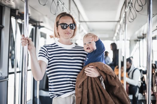 Mother carries her child while standing and holding on to bar holder on bus. Mom holding infant baby boy in her arms while riding in public transportation. Cute toddler traveling with mother