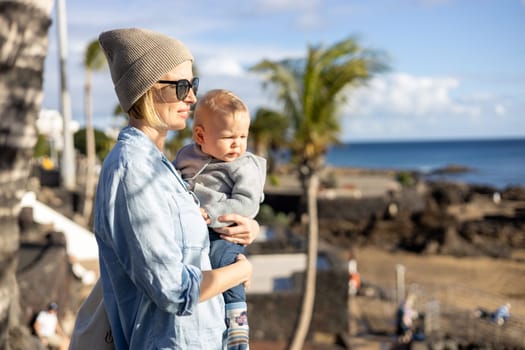 Fashinable mother holding her infant boy child in lap enjoing panoramic view along palm lined beach promenade of Puerto del Carmen, Lanzarote, Canary islands, Spain