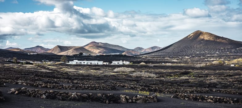 Traditional white houses in black volcanic landscape of La Geria wine growing region with view of Timanfaya National Park in Lanzarote. Touristic attraction in Lanzarote island, Canary Islands, Spain