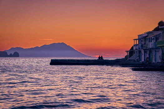 Beautiful view of sunset in Klima village, Milos island, Greece. Pair of adults enjoy sunset view on pier of small fishermen's village. Colorful house, waterfront, distant hazy mountains, calm sea, clear sky, romantic weekend, relax, vacation, voyage