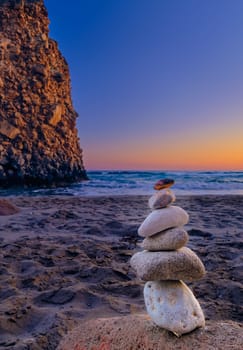 Small balance stone stack and big natural rock lit by sunset sun on sandy beach. Tidal waves in background, selective focus, blurred background, colorful view. Concept of balance, size, meditation, juxtaposition. Vertical shot