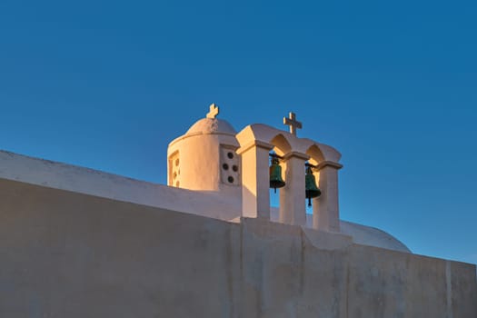Whitewashed Greek Orthodox chapel, small belfry with cross and bell, roof and dome at sunset sky background. Christianity, religion, traditions, symbols, architecture. Close shot. Milos island, Greece