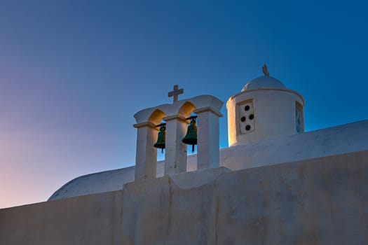 Whitewashed Greek Orthodox chapel, small belfry with cross and bell, roof and dome at sunset sky background. Christianity, religion, traditions, symbols, architecture. Close-up, diagonal shot. Milos island, Greece