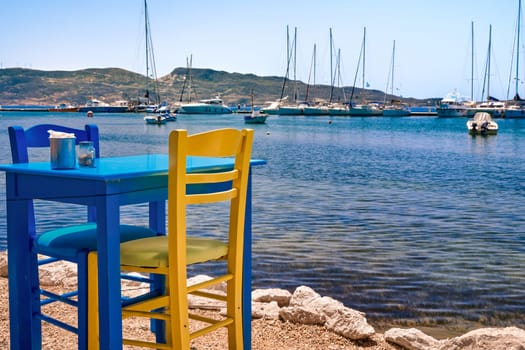 Colorful chair and table of Greek tavern by waterfront at sunny day. Vacations in Greece, summer, restaurant, outdoor dining, port, marina, yachts and boats, Mediterranean relax, Cyclades and Aegean islands. Selective focus, blurred background.