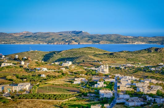Beautiful view of Trypiti town of Milos island, Greece at sunset. Harbour, famous whitewashed houses, green hills and blue sea, distant mountains, clear sky, summer sunshine. Mediterranean style, vacations, romantic weekend, iconic destination