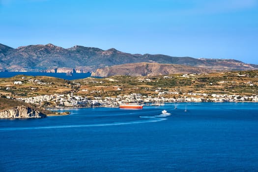 Beautiful summer day and sunshine in typical port town of Greek island. Whitewashed houses by waterfront, ferry moored by pier. Mediterranean lifestyle, vacations, island hopping, cruises. Milos, Cyclades, Greece.