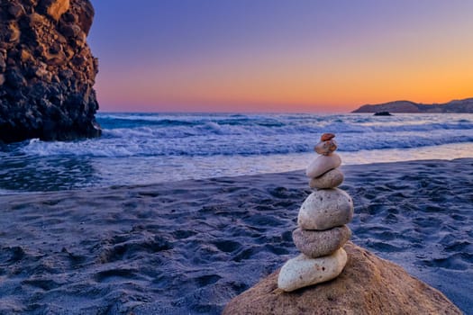 Small balance stone stack and big natural rock lit by sunset sun on sandy beach. Tidal waves in background, selective focus, blurred background, colorful view. Concept of balance, size, meditation, juxtaposition.