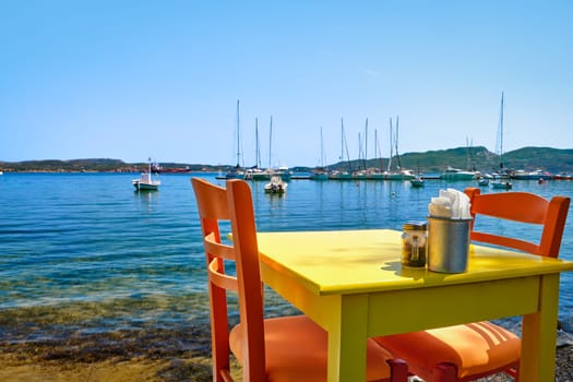 Colorful chairs and tables of Greek tavern by waterfront at sunny day. Vacations in Greece, summer, restaurant, outdoor dining, port, marina, yachts and boats, Mediterranean relax, Cyclades and Aegean islands. Selective focus, blurred background.