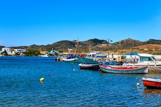 Beautiful summer day and sunshine in typical marina of Greek island village. Colorful fishing boats moored at jetty. Whitewashed houses by waterfront. Mediterranean lifestyle, vacations. Milos, Cyclades, Greece.