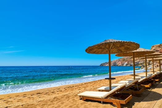 Beautiful empty beach with sun umbrellas and sunbeds. Perfect summer destination. Straw sunshades and sunbeds on the empty beach with blue sea and rocky hills. Vacations in Greece, islands retreat. Milos, Greece.
