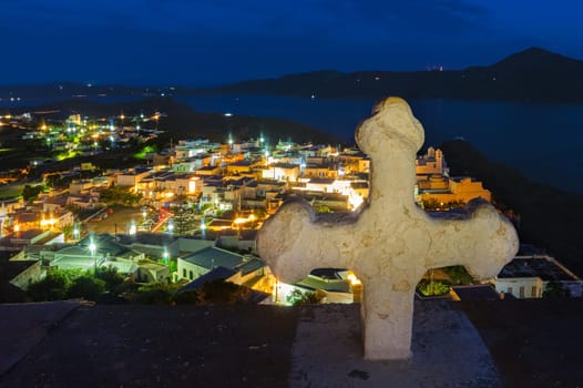 White Greek cross over colorful night view of traditional Greek small town or village by seafront, Plaka, Milos island, Cyclades, Greece. Whitewashed houses, Orthodox church, tiny lit-up streets, distant hills, bay waters, , Mediterranean lifestyle