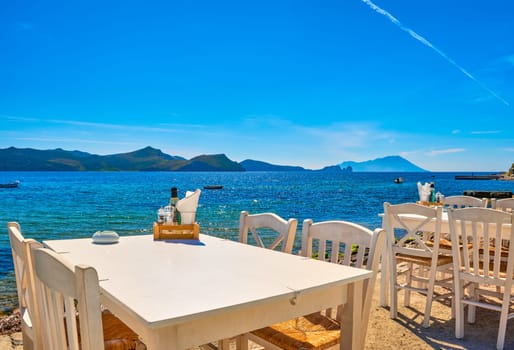 White chairs and tables of Greek tavern by waterfront at sunny day. Vacations in Greece, summer, restaurant, outdoor dining, port, marina, yachts and boats, Mediterranean relax, Cyclades and Aegean islands. Selective focus, blurred background.