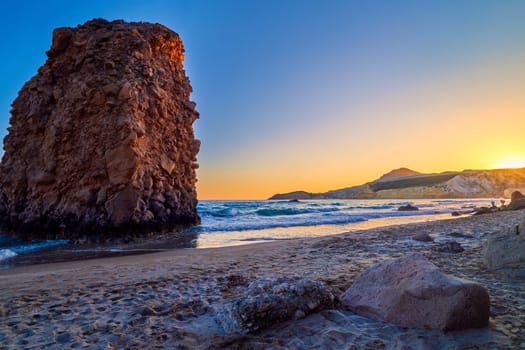 Colorful sunset over Fyriplaka beach, Milos island, Cyclades, Greece. Beautiful sky, unique lonely rock on sandy beach, low sun, sundown haze, rocky hills, sun rays, sea waves spray.