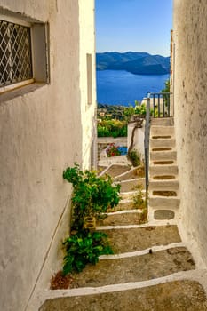 Typical view at sunset in Greek small village by seafront. Whitewashed houses, flower pots, lagoon, vines. Traditional alleyways, summer sunshine, low sun, blue sea waters, distant hills, quiet lifestyle, relax, vacation. Plaka, Milos island, Greece
