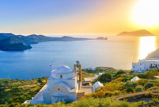 Beautiful view of Mediterranean island at sunset. Whitewashed Greek Orthodox church by seafront in rays of low sun. Sun glare, distant hills, blue sea, clear sky, distant hills, whitewashed houses, summer, vacations, Milos, Cyclades, Greece.