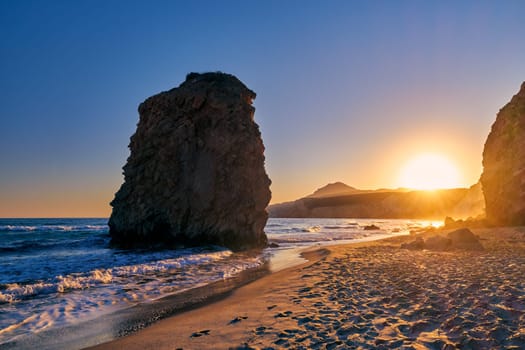 Colorful sunset over Fyriplaka beach, Milos island, Cyclades, Greece. Beautiful sky, unique lonely rock on sandy beach, low sun, sundown haze, rocky hills, sun rays, sea waves spray.