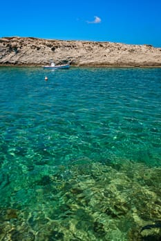 Anchored small boat under Greek flag, azure sea waters, clear blue sky, rocky hill, red fishing buoy floating. Frontal shot. Milos, Greece. Summer vacations background, concept, copy space, no people, vertical composition, book cover.