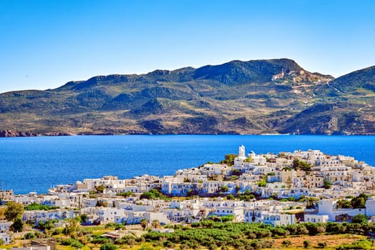 Beautiful view of Adamantas, main town of Milos island, Greece, on sunny day. Hill top town, whitewashed houses, church bell tower, blue sea waters of bay and distant hazy hills. Greek islands, Mediterranean sea, summer sunshine.