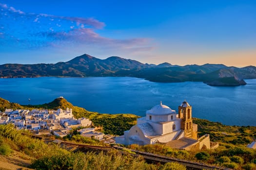 Beautiful view of Mediterranean island at sunset. Whitewashed Greek Orthodox church by seafront in rays of low sun. Sun glare, distant hills, blue sea, clear sky, distant hills, whitewashed houses, summer, vacations, Milos, Cyclades, Greece.