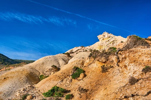 Unique natural rock formations of yellow and orange cliffs in low sunlight, Milos, Greece. Weather-shaped slopes of rocks on Agios Ioannis beach at clear blue sky.