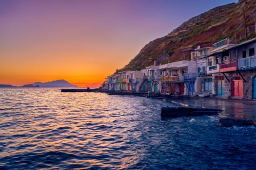 Beautiful view of sunset in Klima village, Milos island, Greece. Pair of adults enjoy sunset view on pier of small fishermen's village. Colorful houses by waterfront, distant mountains, calm sea, clear sky, romantic weekend, relax, vacations, voyage