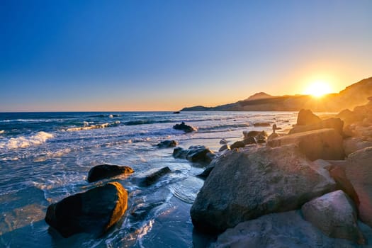 Colorful sunset over Fyriplaka beach, Milos island, Cyclades, Greece. Beautiful sky, low sun, sundown haze, rocky hills, sun rays, sea waves spray beating on natural rock formations.