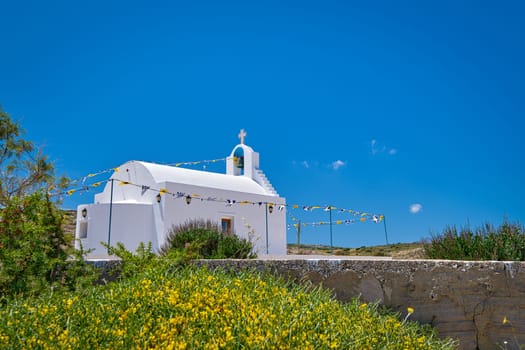 White Greek Orthodox chapel or church on hilltop of seashore against clear blue sky on sunny day. Traditional religious building. Colorful flowers and bushes in foreground. Typical Greek landscape. Diagonal composition, copyspace. Milos Greece