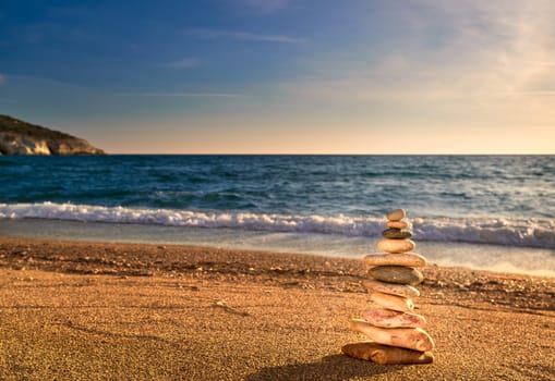Small balance stone stack or cairn on sandy beach in sunset sunlight, sea waves. . Tidal waves in background, selective focus, blurred background, colorful view. Concept of balance, meditation, peach and zen.