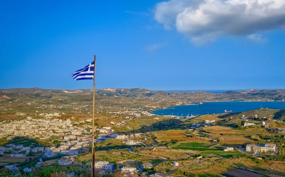 National flag of Greece waving over landscape of Milos island. Adamantas town, harbour and villages at sunset. Famous whitewashed houses, green hills and blue sea, distant mountains, clear sky with cloud, summer sunshine. Mediterranean vacations