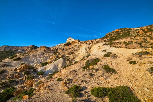 Unique natural rock formations of yellow and orange cliffs in low sunlight, Milos, Greece. Weather-shaped slopes of rocks on Agios Ioannis beach at clear blue sky.