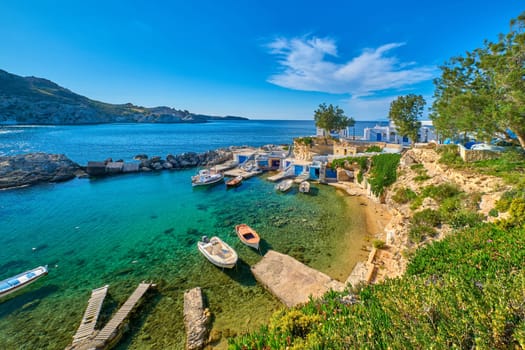 Beautiful view of secluded Mediterranean island harbour on summer day. Whitewashed houses, hills, clear blue sky, azure sea waters, beach, anchored small motor boats. Milos island, Cyclades, Greece. Mediterranean getaway vacations on remote islands.