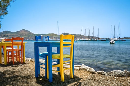 Colorful chairs and tables of Greek tavern by waterfront at sunny day. Vacations in Greece, summer, restaurant, outdoor dining, port, marina, yachts and boats, Mediterranean relax, Cyclades and Aegean islands. Selective focus, blurred background.