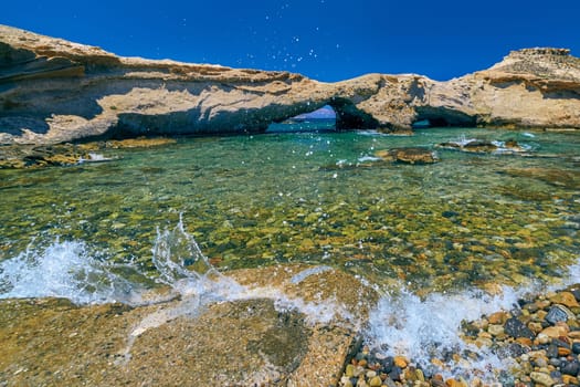 Tide splashes on rocky shore, small bay with azure clear sea waters, islet caves, stone arches. Mediterranean vacation, summer holiday, sunny summer day, swimming. Great blue sky, no clouds, rocky cliffs, little greenery, island. Milos island, Greece