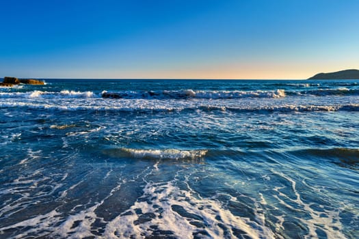 Beautiful seascape of low tidal waves, clear sunset sky, hazy hills. Clear blue and azure waters approaching with little white foam. Nature, landscape, calm and relaxing meditative view. Mediterranean sea, Greece.