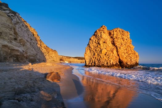 Colorful sunset over Fyriplaka beach, Milos island, Cyclades, Greece. Unique lonely rock on sandy beach lit by low sun mirroring in wet sand of tide. Beautiful sky, rocky hills, sea waves spray, no people.