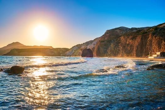 Beautiful landscape of Fyriplaka beach, Milos, Greece. Low sun over azure sea waters, rocky shores, sandy beach, hazy distant rocks. Colorful sun and sky, sun flares on waves.