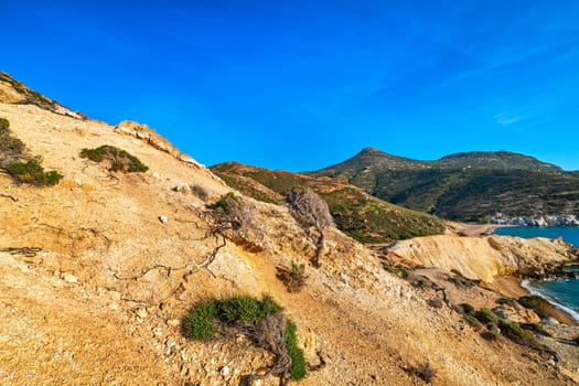 Unique natural rock formations of yellow and orange cliffs in low sunlight, Milos, Greece. Weather-shaped slopes of rocks on Agios Ioannis beach at clear blue sky.