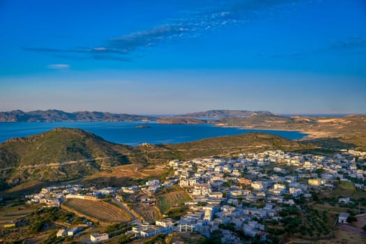 Beautiful view of typical Greek town on island, Greece at sunset. Harbour, famous whitewashed houses, green hills and blue sea, distant mountains, clear sky, summer sunshine. Mediterranean style, vacations, romantic weekend, iconic destinations